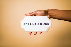Close-up of a hand holding a 'Buy Our Giftcard' sign on a brown backdrop.